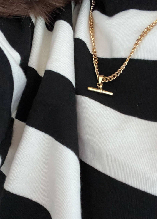 Gold necklace with a T-bar pendant on a black and white striped fabric background