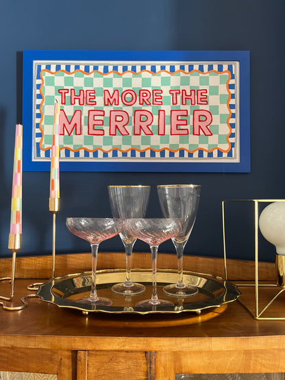 A textile wallhanging with a blue frame and orange and turquoise borders, featuring the phrase 'THE MORE THE MERRIER' on a turquoise checkered fabric displayed above a wooden table with glass glasses and decorative items.