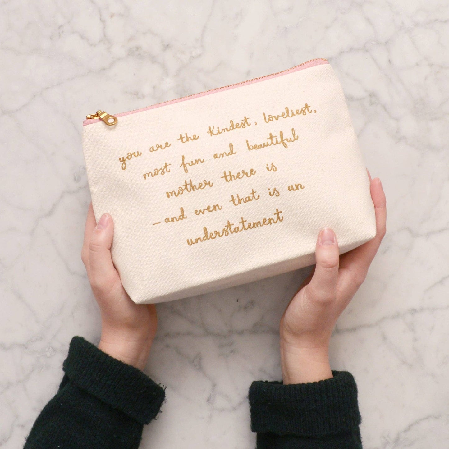 Cream-colored pouch with gold text held by hands on a marble surface