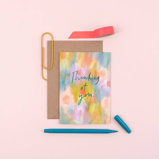 Greeting card with 'Thinking of you' text on a colorful watercolor background, with a pen and paper clips on a pink background.