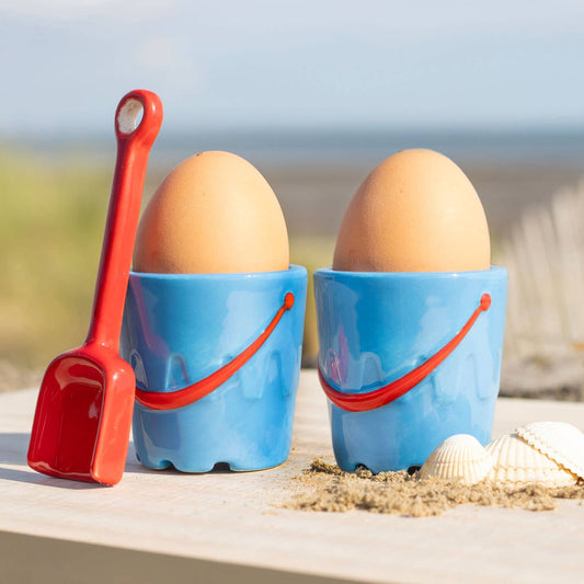 Two eggs in blue sand pails with red shovels on a beach.