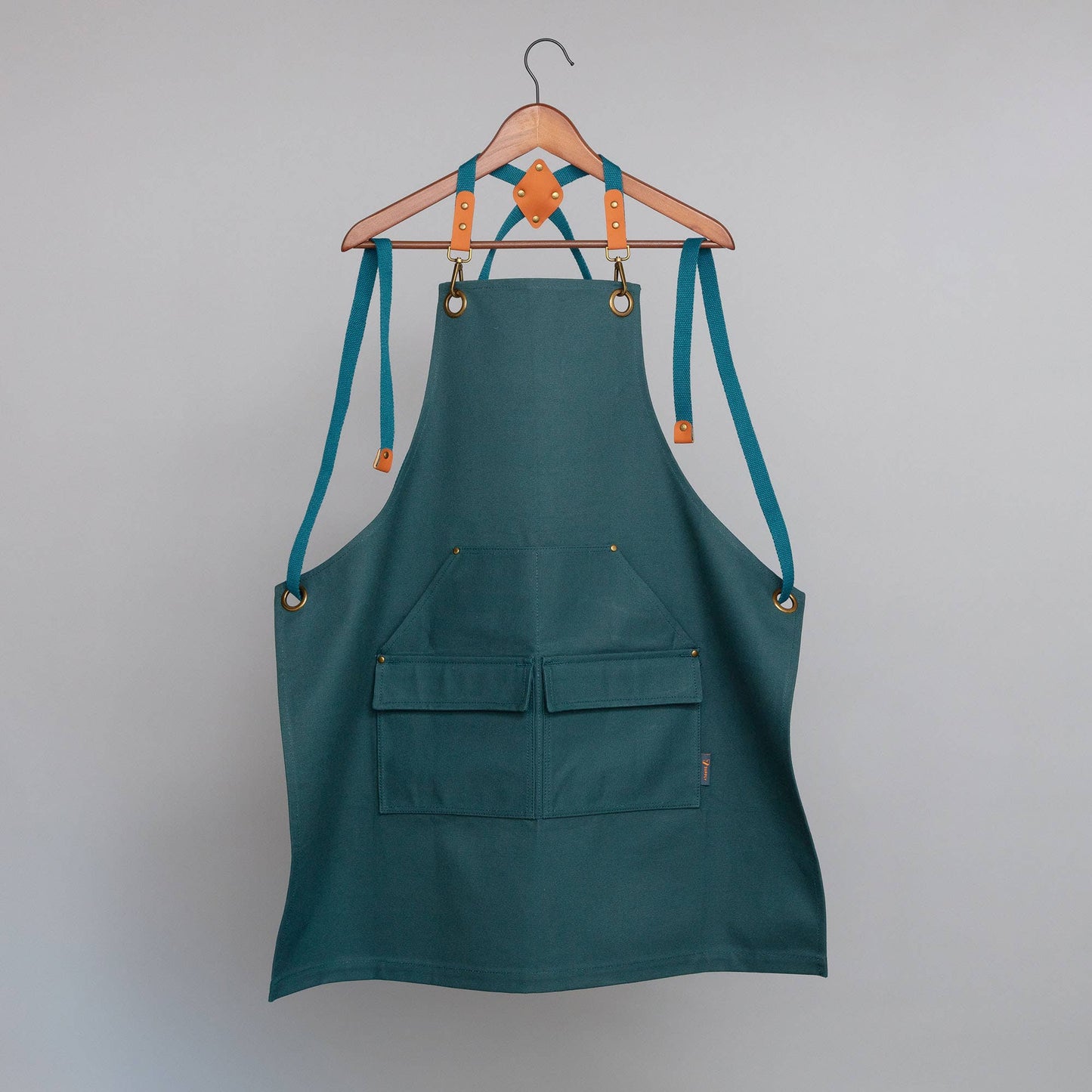 Green apron with teal straps hanging on a wooden hanger against a gray background