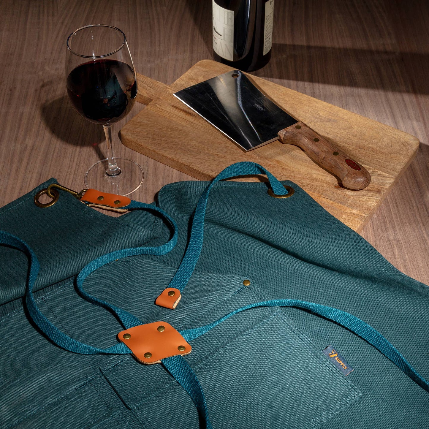 Teal apron with orange accents on a wooden table with a glass of red wine and a knife.