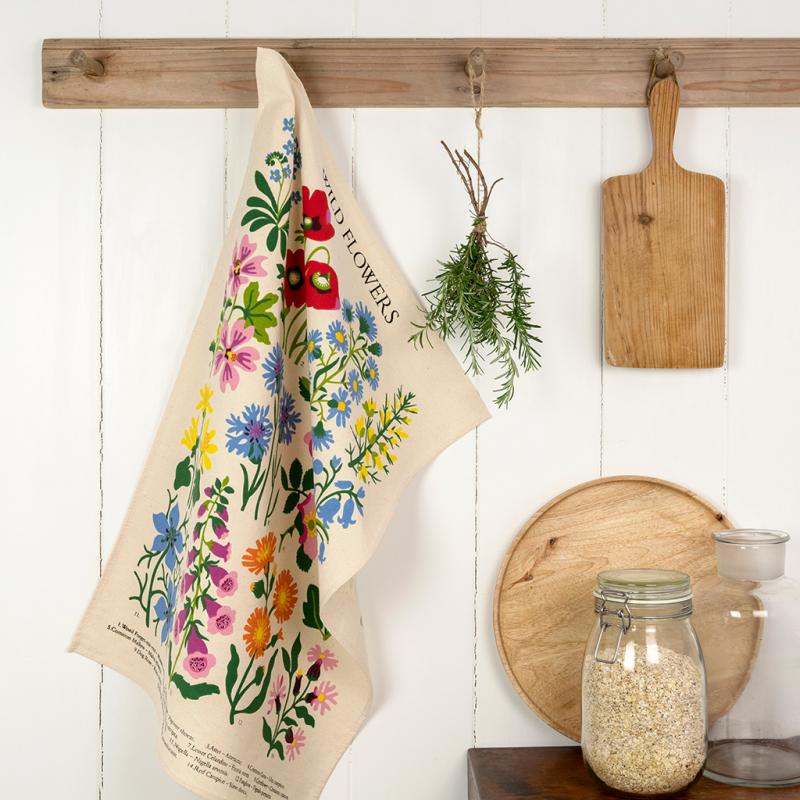 A multicolored floral print cotton tea towel hanging on a wooden rack in a kitchen setting, with a cutting board and some herbs in the background.