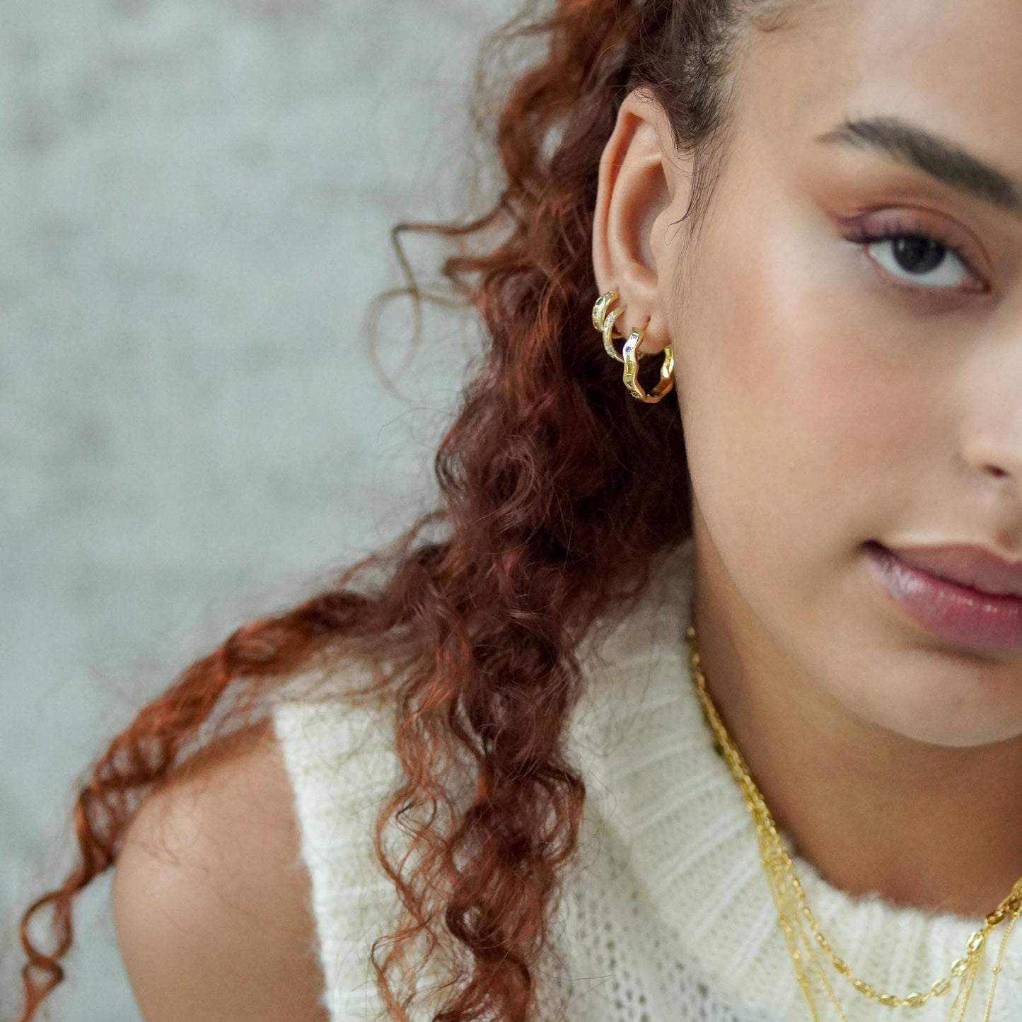 Close-up of a woman wearing gold hoop earrings and necklaces against a neutral background